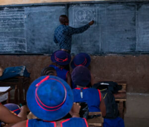 A classroom where a teacher in a blue t-shirt is drawing diagrams on the blackboard. Pupils dressed in blue uniforms are sitting at their desks and looking at him with attention.