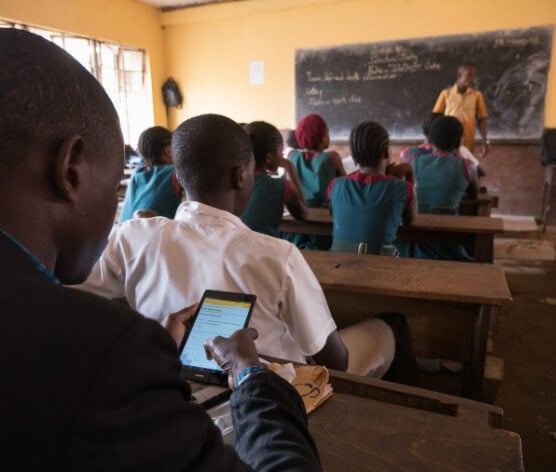 A classroom with a teacher doing a presentation to his pupils. In the first rows, some pupils dressed in dark green uniforms are paying attention. At the back, a supervisor is using a tablet to record monitoring data.