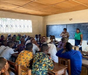 A classroom with two school leaders talking in front of the blackboard to several boys sitting on the benches and listening carefully. 