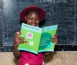 A girl, wearing a red uniform and hat, is reading happily a green textbook in front of the blackboard.