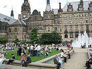 View across a garden containing people enjoying a sunny day towards a large Victorian building with a clock tower