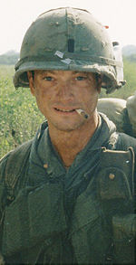 A man is at the center of the image looking at the camera. He is dressed in Vietnam-era military attire including a vest and helmet. He has a cigarette sitting on his lips and is wearing a backpack.