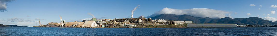 A panorama featuring a large industrial plant on a sea side, in front of mountains.