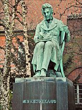 A statue. The figure is depicted as sitting and writing, with a book on his lap open. Trees and red tiled roof is in background. The statue itself is mostly green, with streaks of grey showing wear and tear. The statue's base is grey and reads 