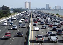 Looking down a straight busy ten-lane highway running along a coastline. In the background are a few multi-story buildings.