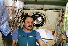 A man, dressed in blue work clothing, seen in a small cubicle. On the walls around him can be seen a sleeping bag, children's drawings, technical manuals and stained insulation. A small porthole in the centre of the wall behind him shows the nose of the Space Shuttle Atlantis and the blackness of space.