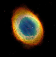 Elliptical shell with fine red outer edge surrounding region of yellow and then pink around a nearly circular blue area with the central star at its center. A few background stars are visible.