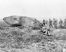 Files of soldiers with rifles slung follow close behind a tank, there is a dead body in the foreground