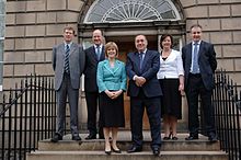 Four men and two women in smart dress pose on steps to large town building.