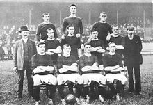 A black-and-white photograph of a football team lining up before a match. Four players, wearing dark shirts, light shorts and dark socks, are seated. Four more players are standing immediately behind them, and three more are standing on a higher level on the back row. Two men in suits are standing on either side of the players.