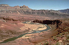 Layered dark brown rock in stairstep pattern in ledges above a river in a canyon with exposed reddish and tan rock