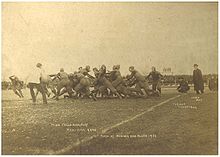 A grainy black and white photo of teams in sports jerseys and light padding contesting a play near the touchline, while watched by officials, coaches and crowds.