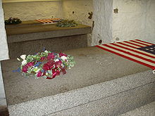 3 marble sarcophagi, one in the foreground, 2 in the background are seen. 2 are seen with flags of the United States at the top.