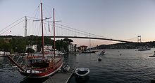 A docked sailboat floats in front of a suspension bridge, under twilight.