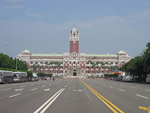 A tall and large building with a tower in its center. A large road surrounded by trees leads to it.