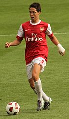 A young black man stands on a pitch dressed in football kit. The body of his shirt is red and bears the club badge and logos of manufacturer and sponsor on the front. The rest of his kit, long sleeves, shorts, socks and boots, is white. His shorts bear the number 32.